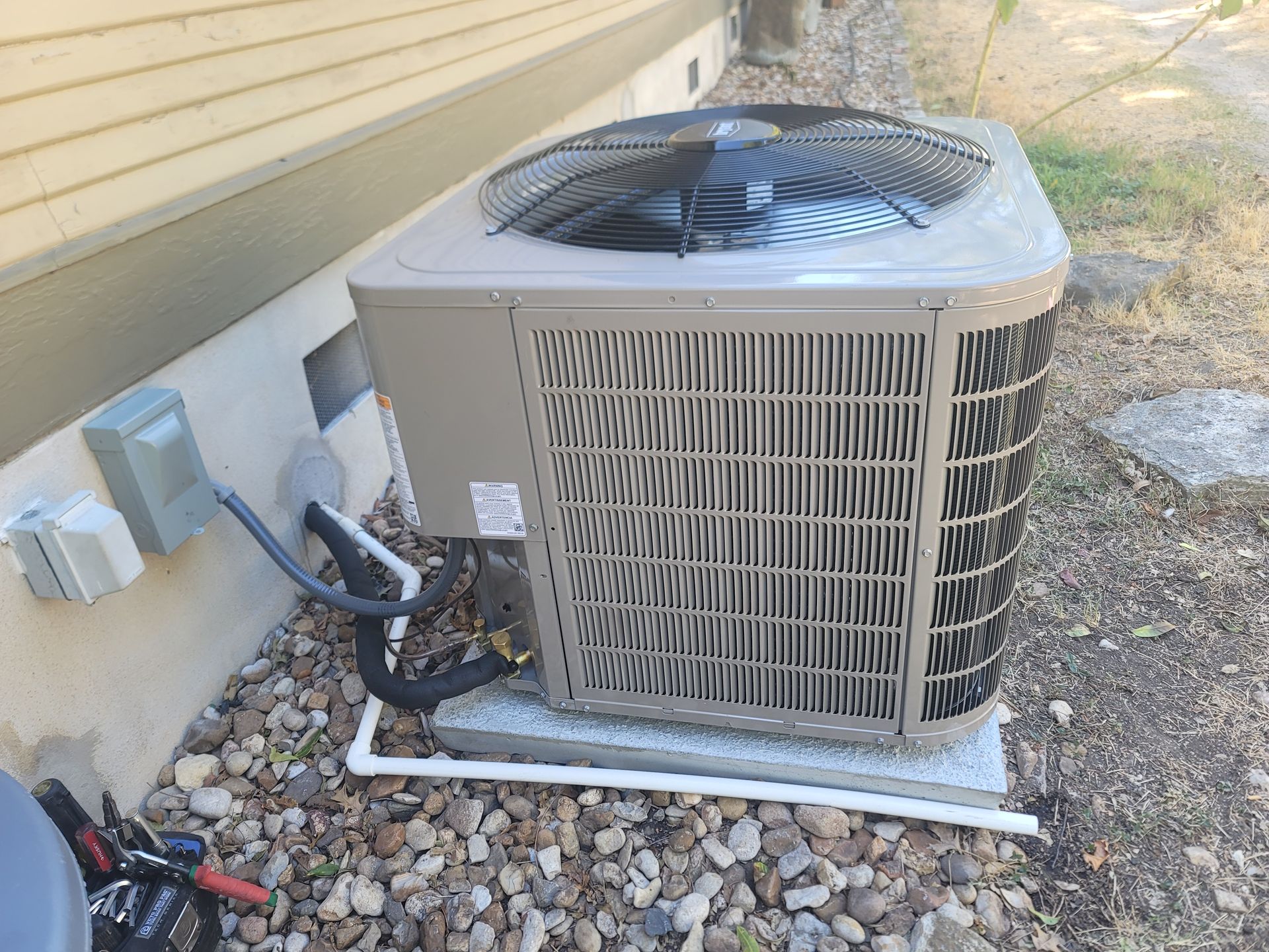 Outdoor air conditioner unit beside a house wall, with rocks and electrical boxes nearby.