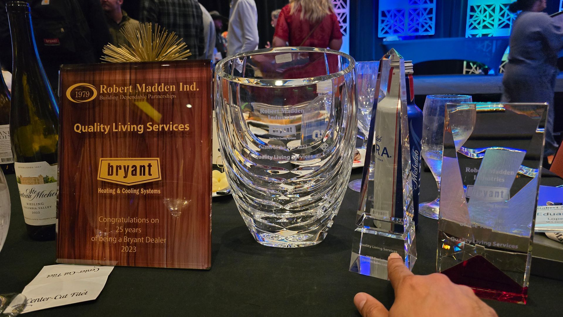 Award plaques on a table, with a hand pointing at a crystal trophy in a dimly lit event setting
