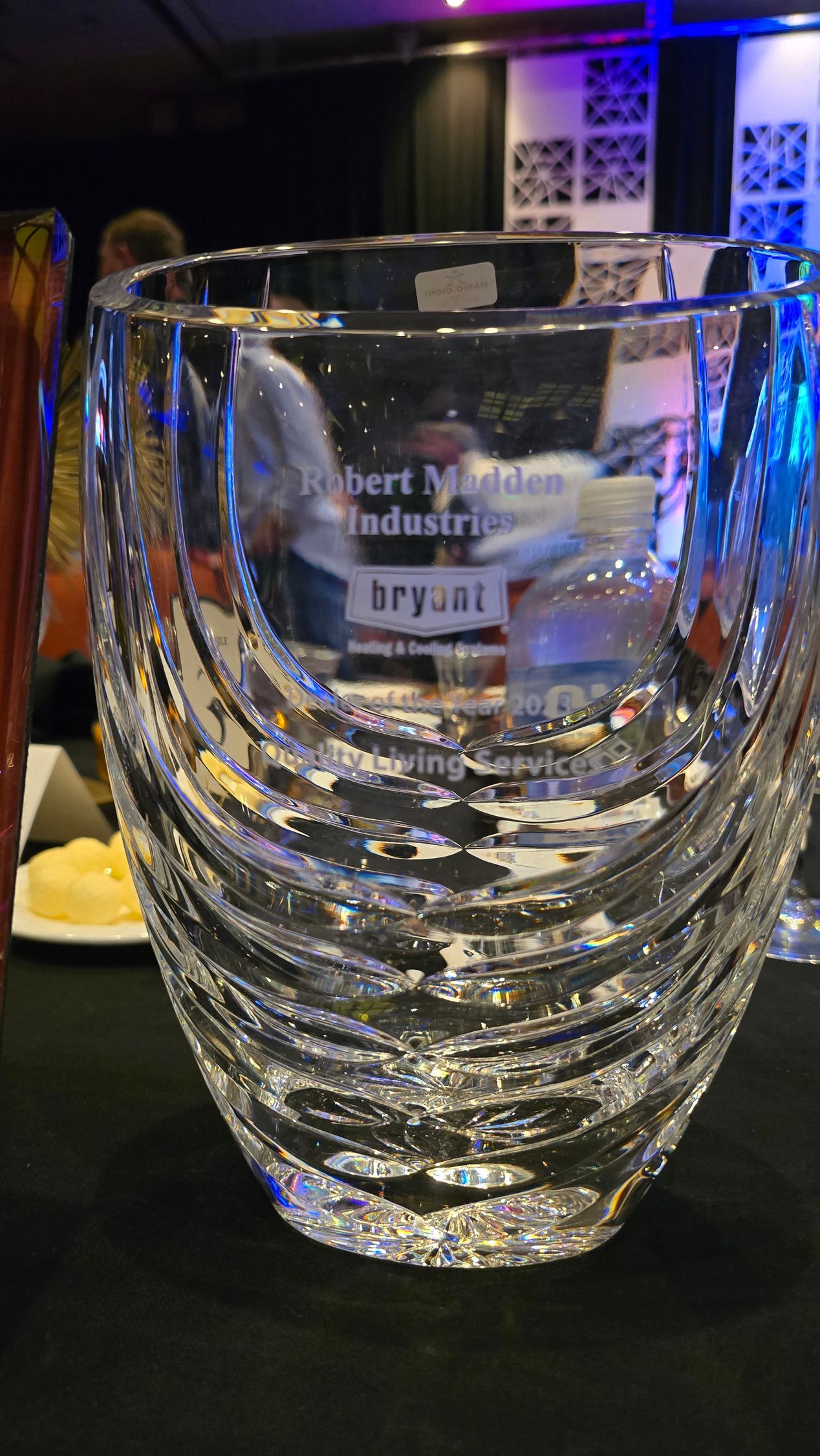 Clear etched glass award on a dark table, with a colorful lit background and other plaques behind it