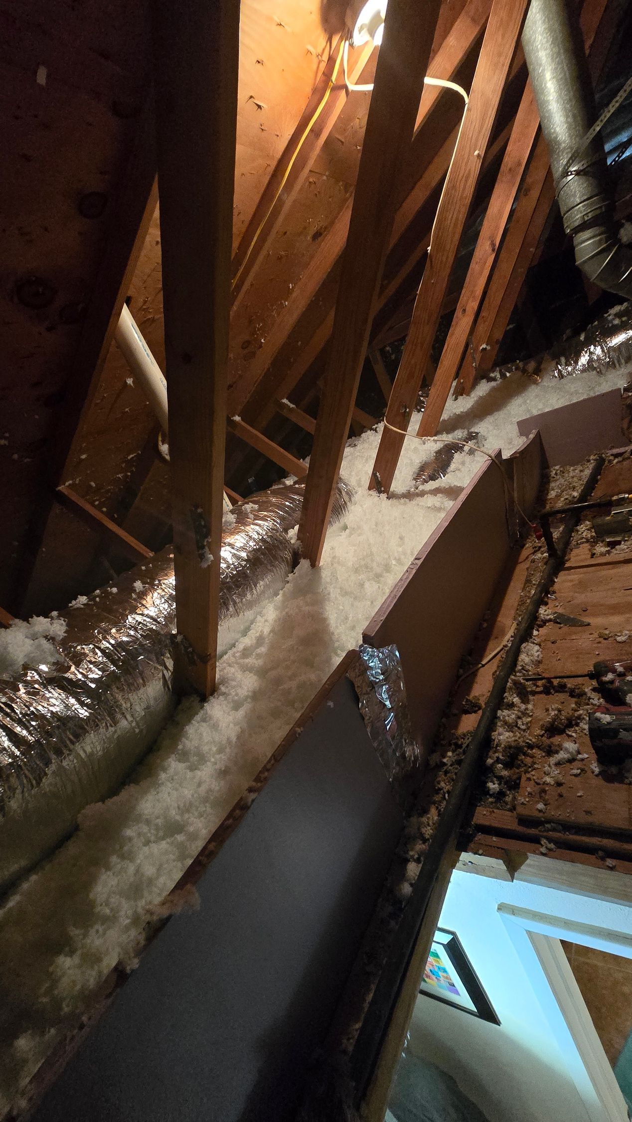 Narrow attic crawlspace with exposed wooden beams, insulation, and a dark walkway under a slanted roof.