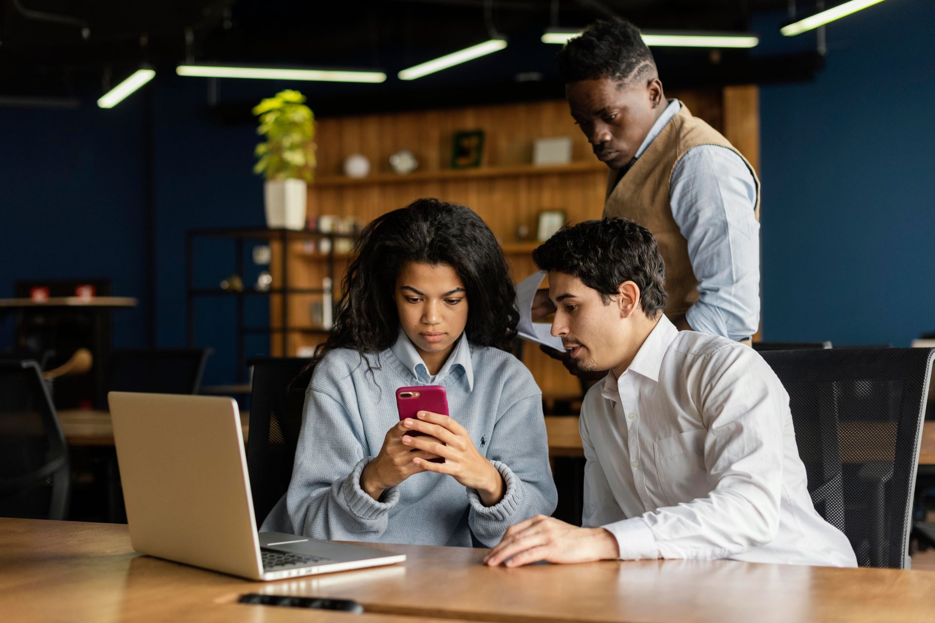 Três colegas trabalham juntos em uma mesa, olhando para a tela de um smartphone em um escritório moderno