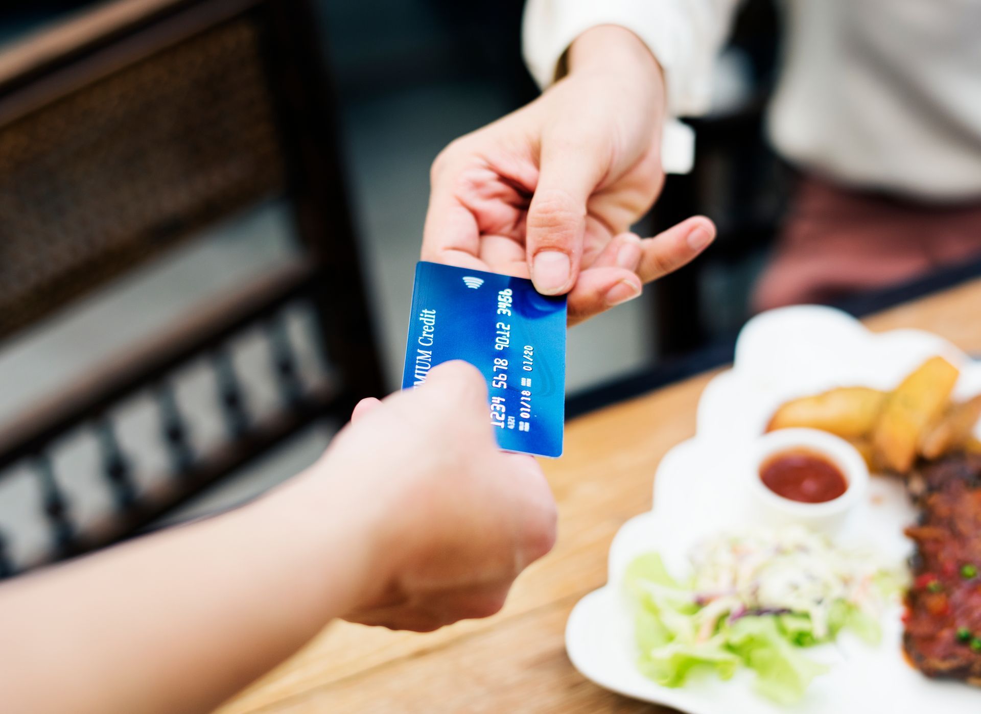 Duas pessoas trocando um cartão de crédito azul à mesa, durante uma refeição em um restaurante.