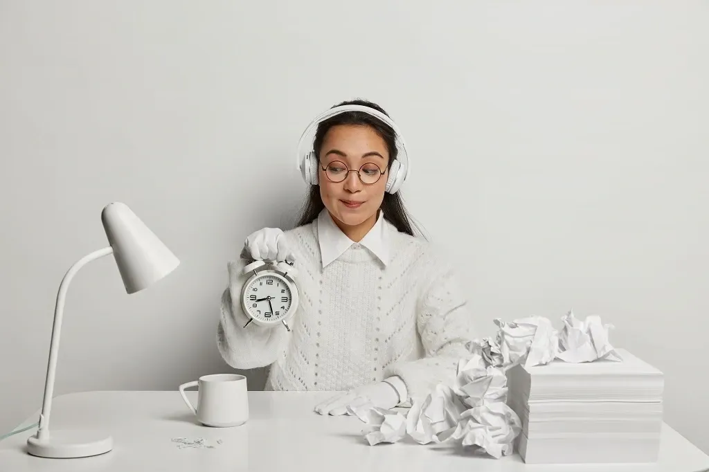 Mulher segurando um despertador, cercada por papéis amassados. Usa fones de ouvido, óculos e um suéter.