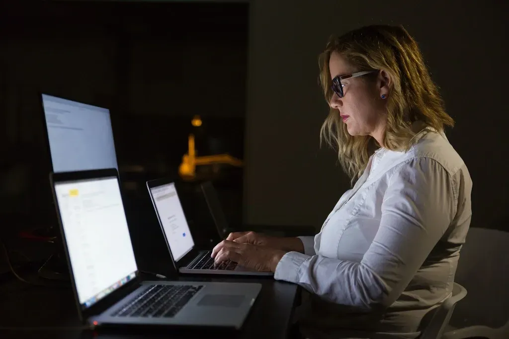 uma mulher de cabelo loiro e curto vestindo camisa branca e óculos está de frente para computadores a noite
