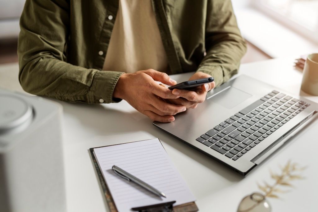Uma pessoa sentada em umacadeira de frente para uma mesa com notebook usa o celular com as mãos