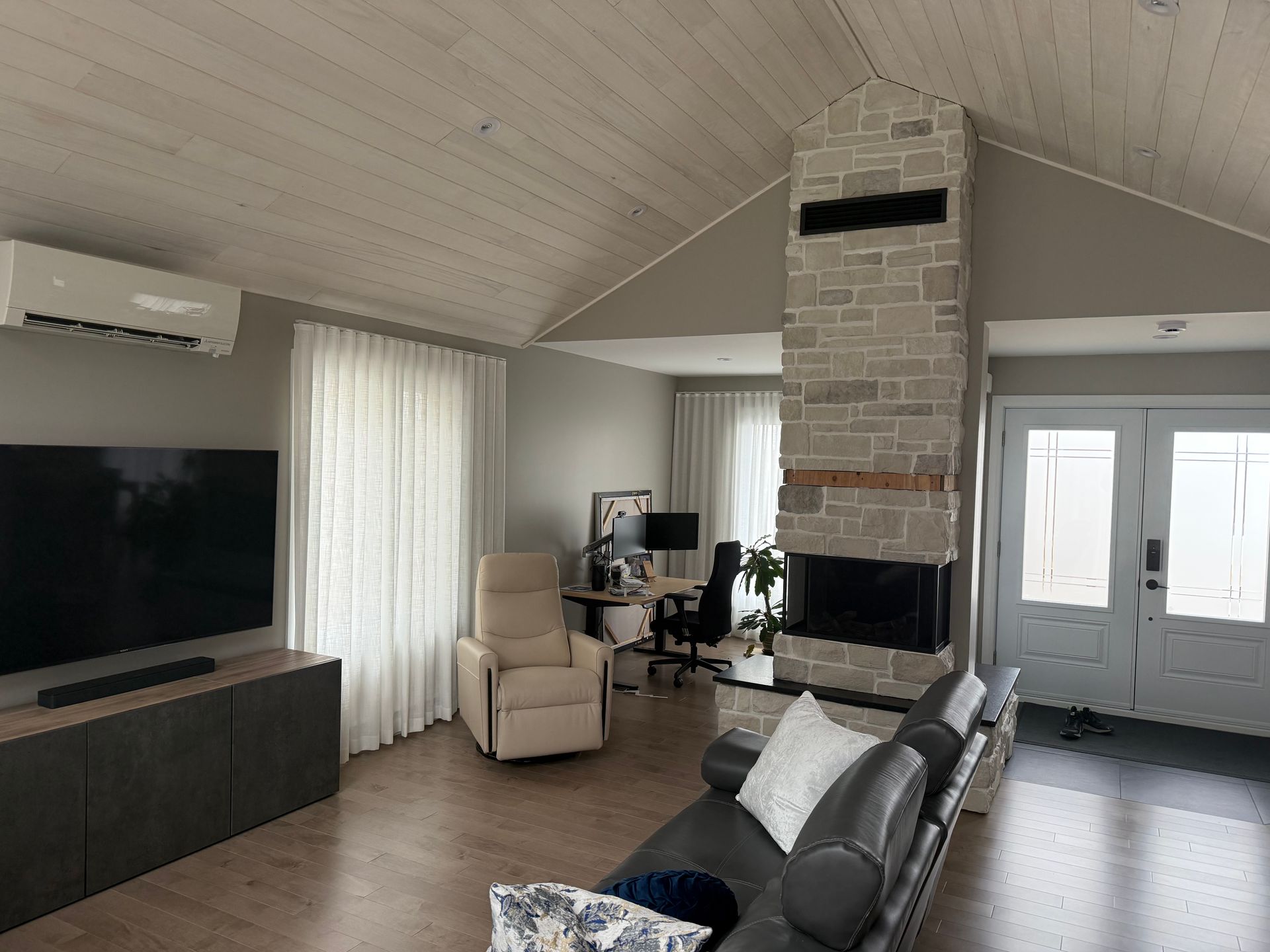 Salon lumineux avec cheminée, télévision, fauteuil beige, canapé gris et portes blanches sous un plafond voûté