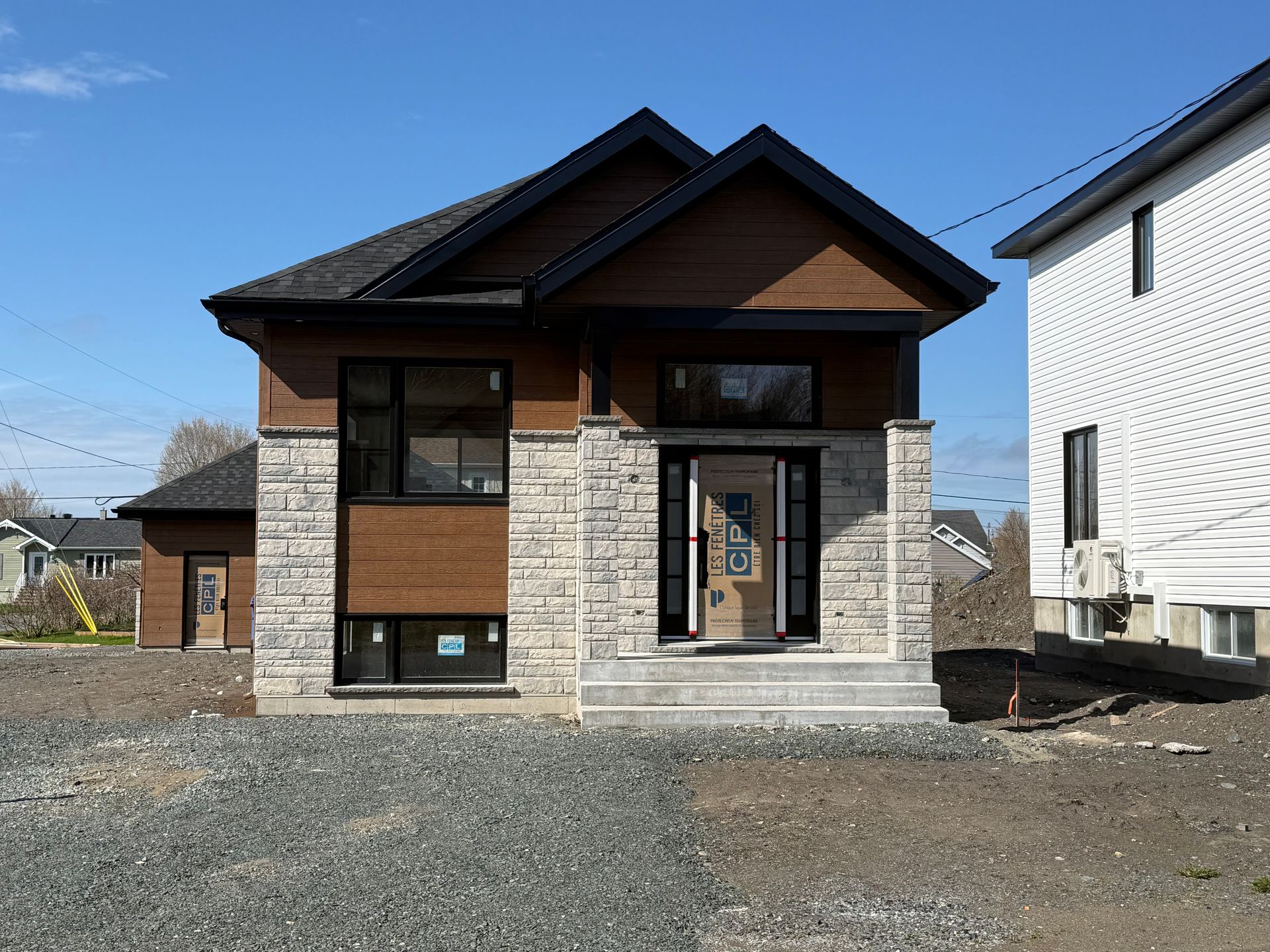 Maison neuve de deux étages avec extérieur en pierre et en bois sur un terrain de gravier sous un ciel bleu clair