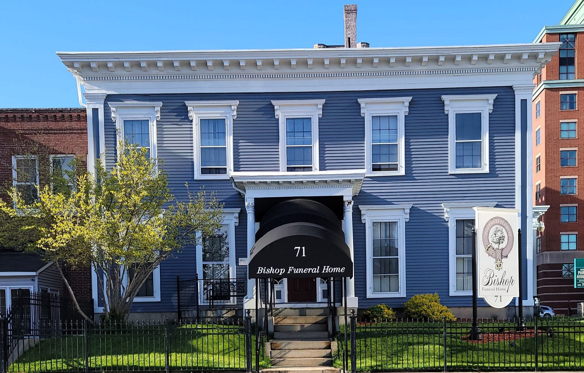 Blue historic house with white trim and a front awning behind a black fence on a sunny day