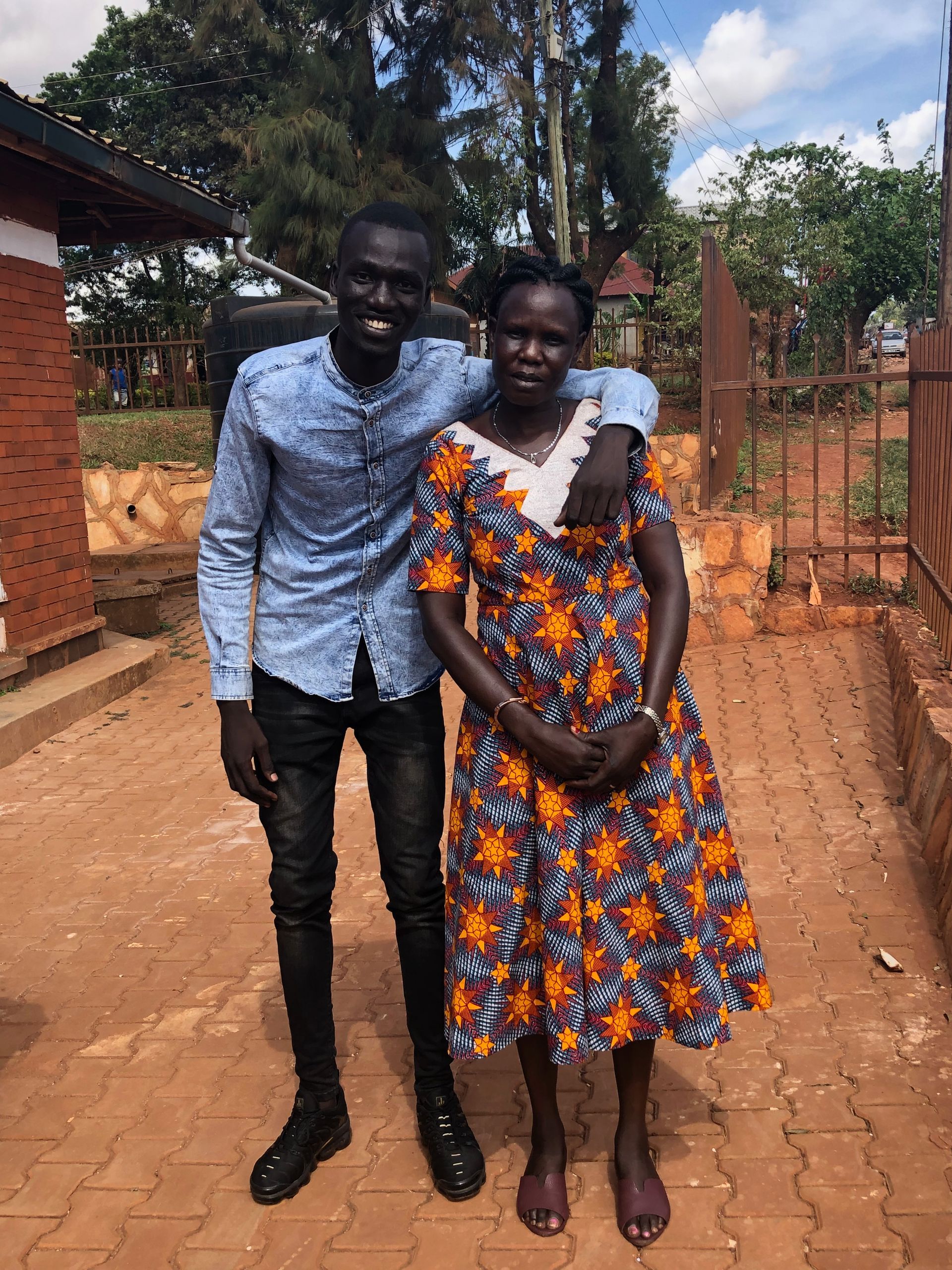 Man with arm around woman, smiling, in front of a brick building. The woman wears a patterned dress.