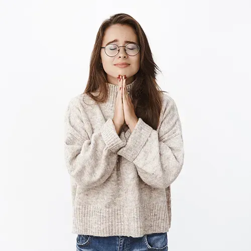 Woman with glasses wearing a light sweater and jeans, hands clasped in front, eyes closed.