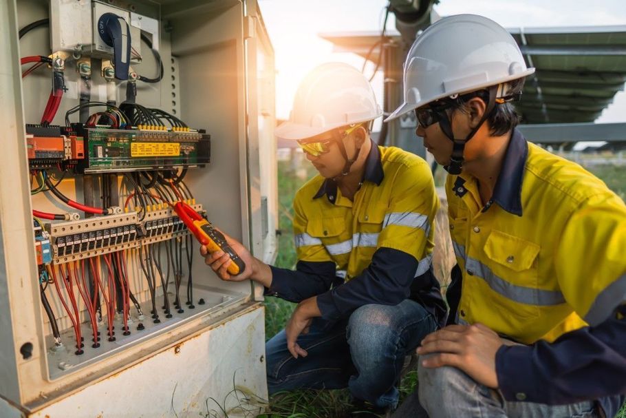 Two workers in hard hats inspect an electrical panel outdoors at sunset.