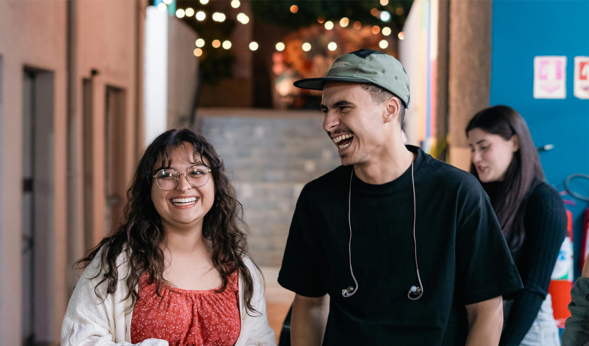 Two smiling people walking indoors, one in a red dress and one in a black shirt and cap, with string lights behind them