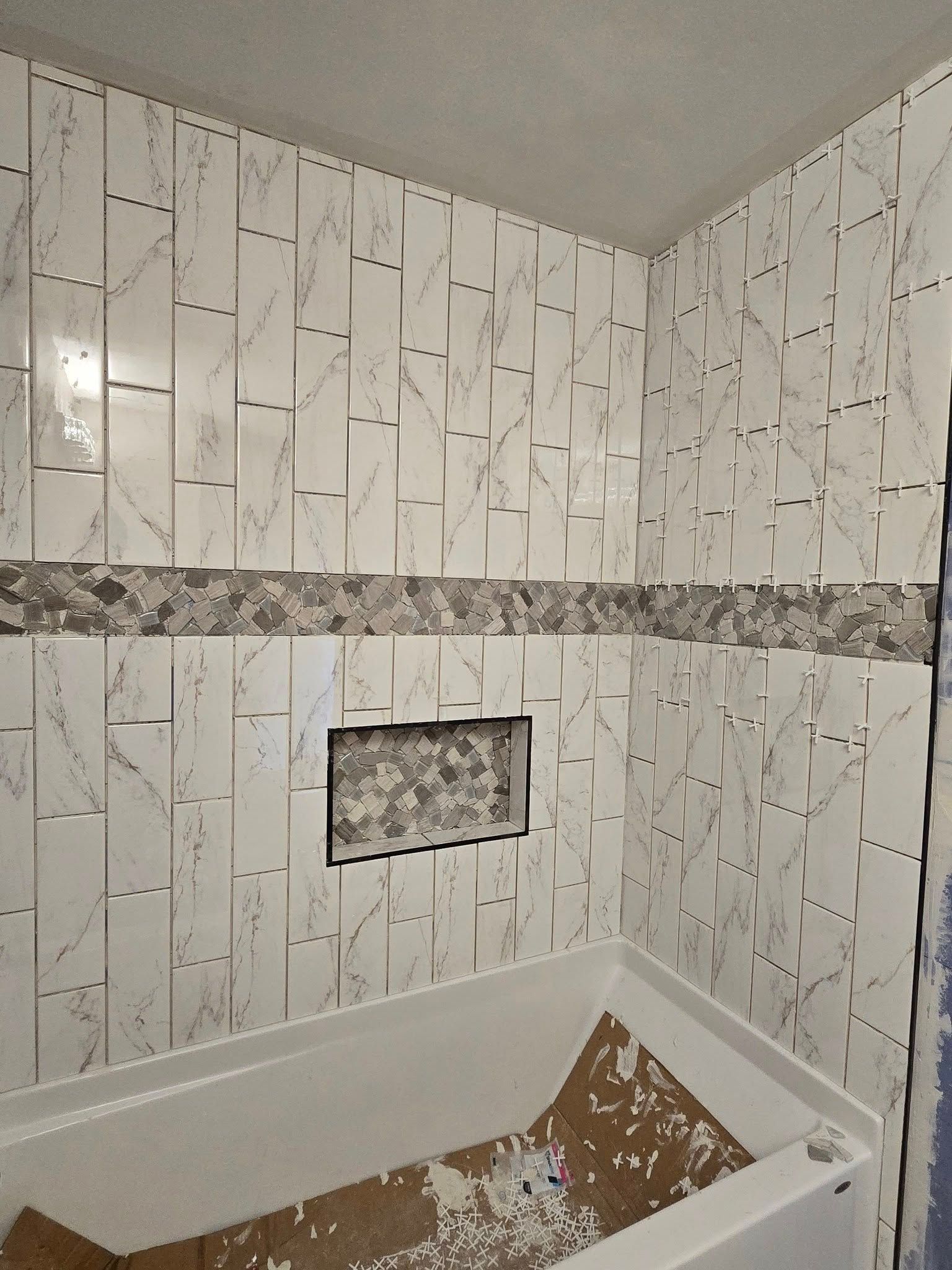 Partially tiled bathroom shower with white marble-look walls and an unfinished bathtub below the tile line