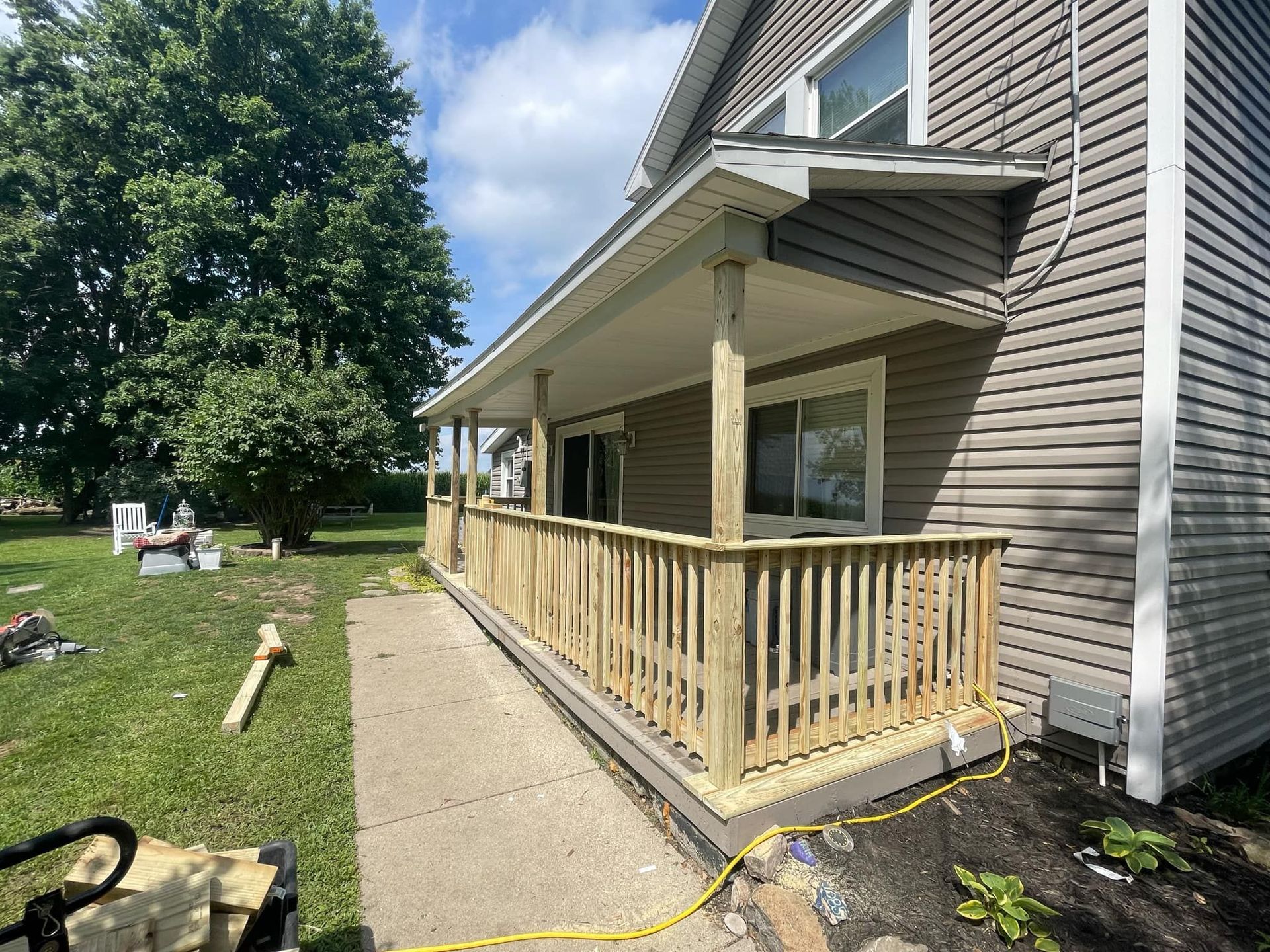 Wooden porch under construction beside a gray house, with railing, gravel path, and yellow extension cord