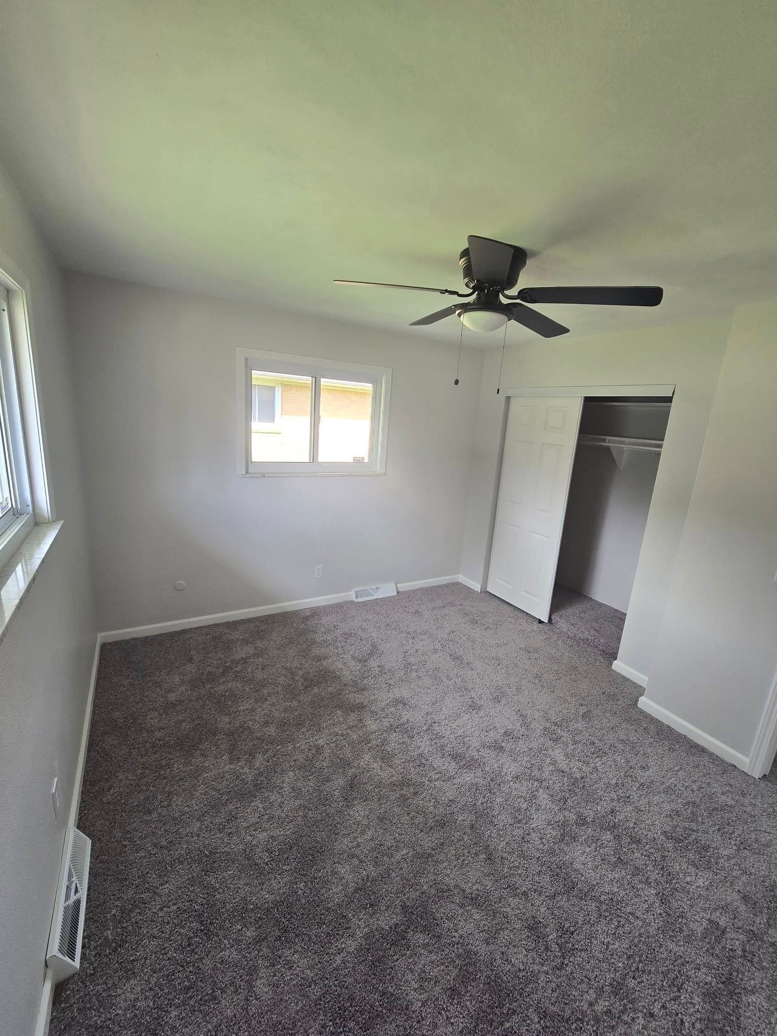 Empty gray bedroom with carpet, ceiling fan, two windows, and an open closet door.