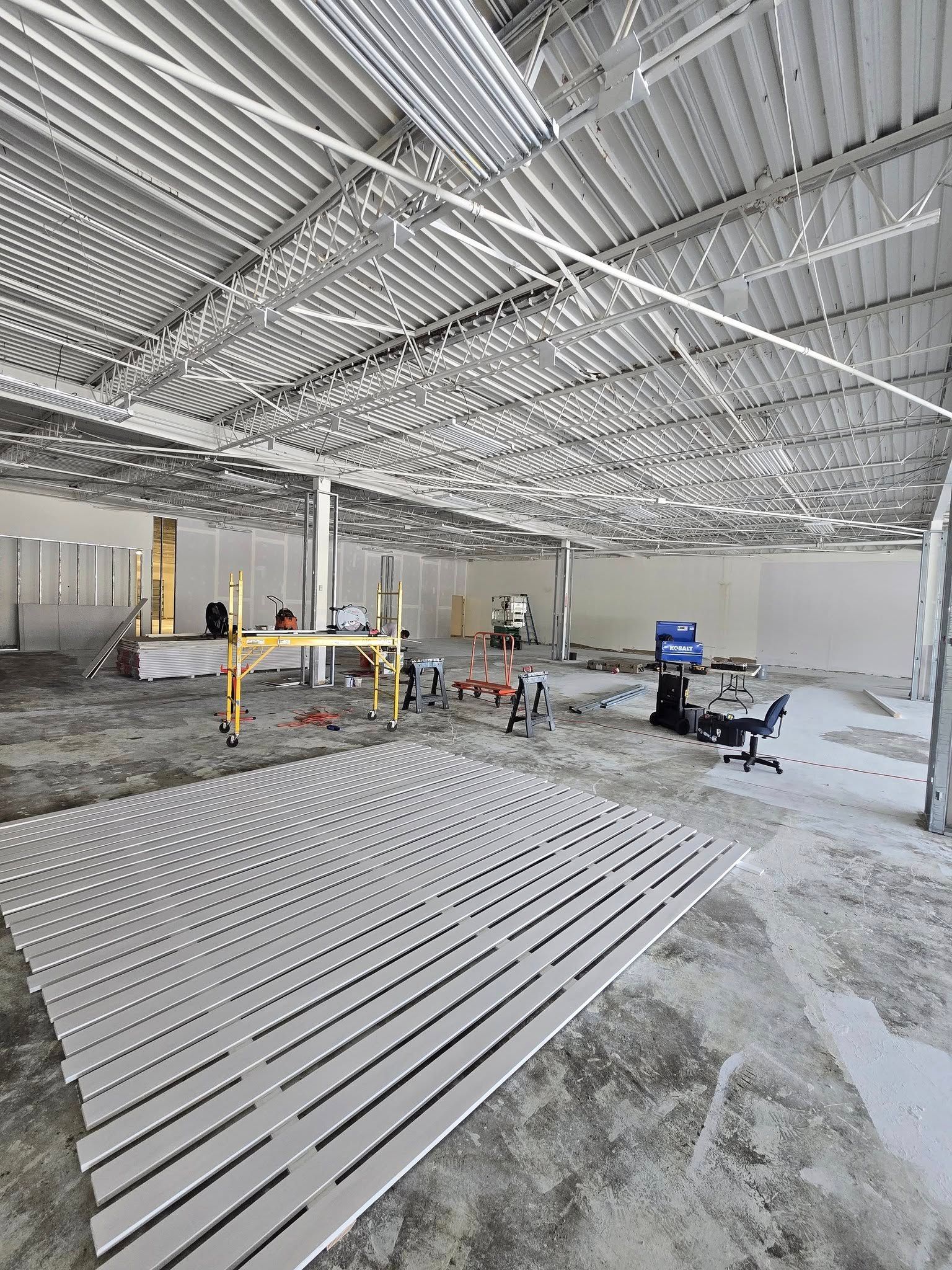 Construction site with metal ceiling grid and workers amid stacked white slats and equipment