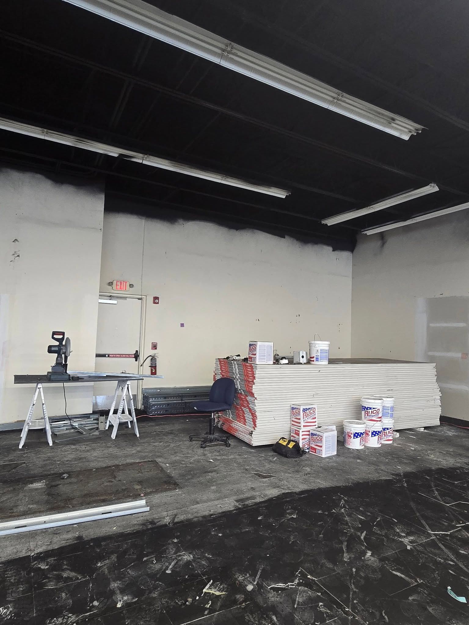 Empty warehouse interior with stacked materials, worktable, and tools under fluorescent ceiling lights