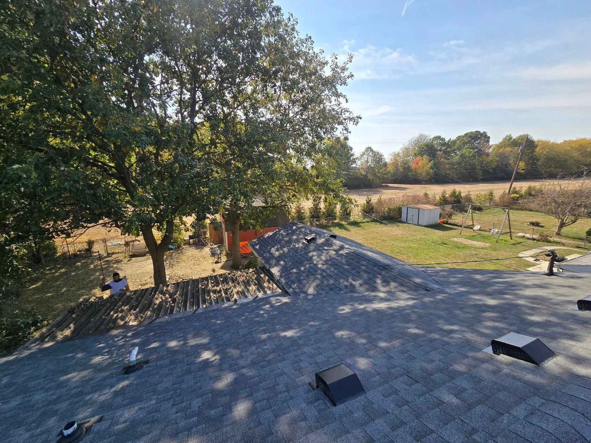 Gravel rooftop under construction beside a tree, with fields and a clear sky in the background
