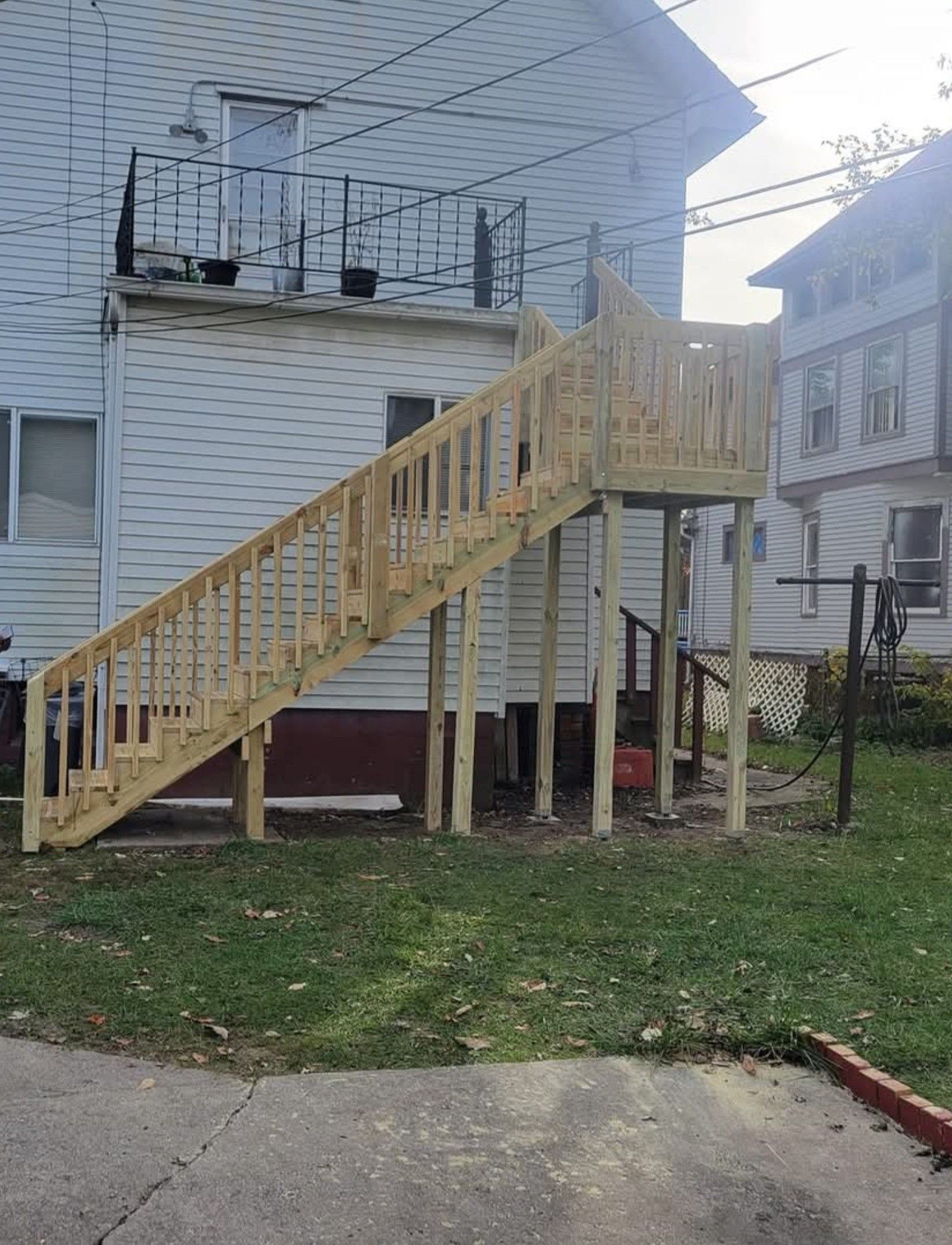Wooden outdoor staircase leading to a raised white house, with railings and posts in a backyard.