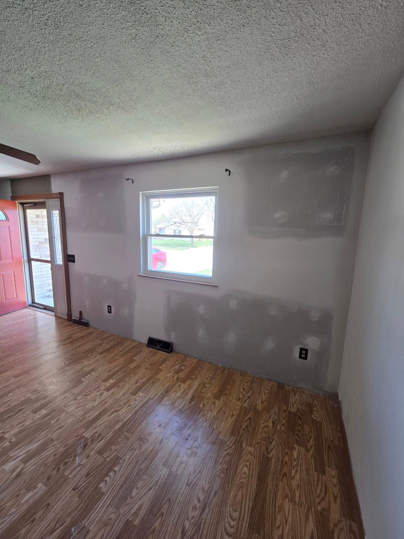 Empty living room with wood floors, white walls, a window, and an open doorway to a red exterior area.