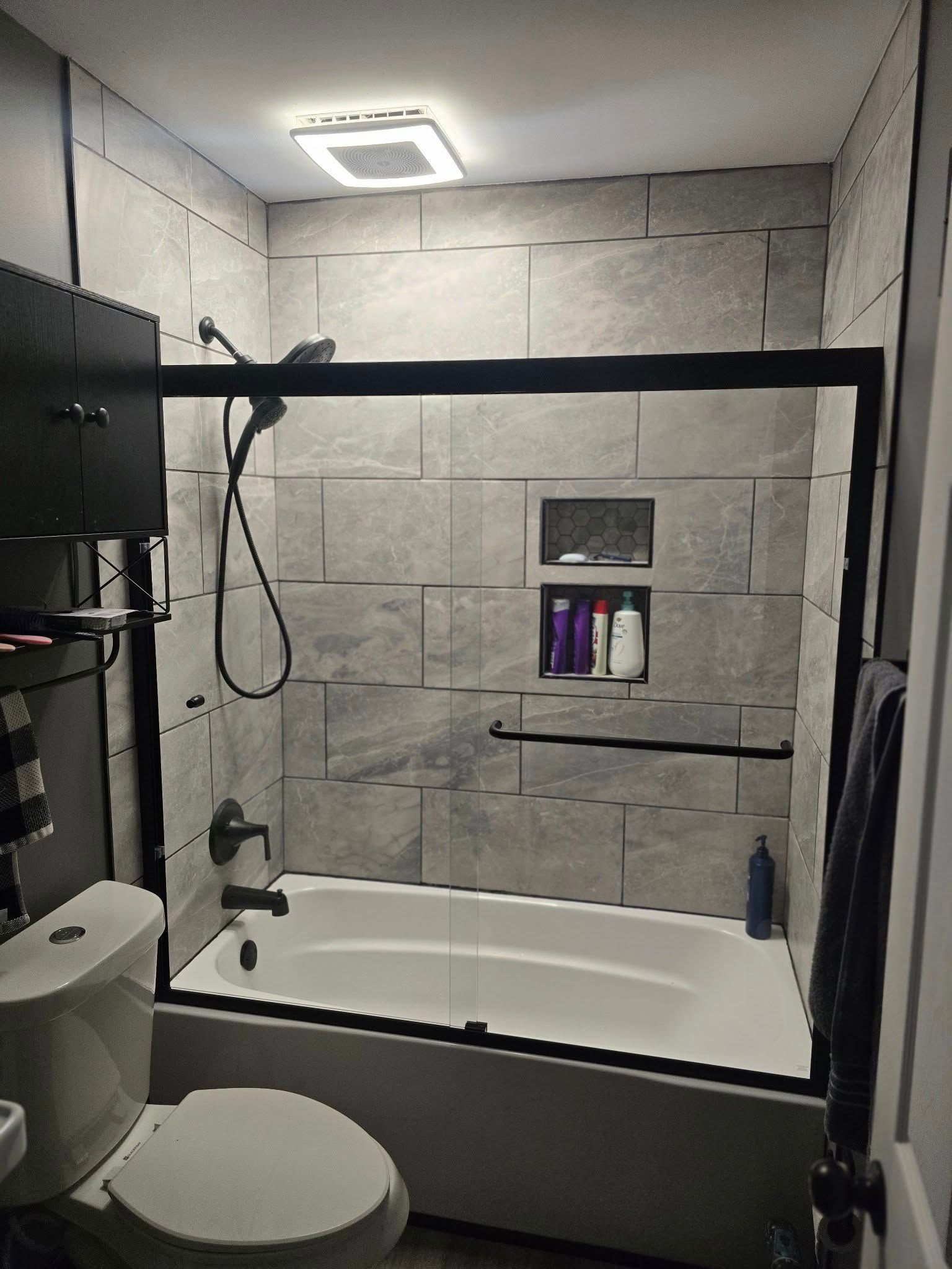Small bathroom with gray tiled walls, bathtub, glass shower enclosure, toilet, and ceiling light.
