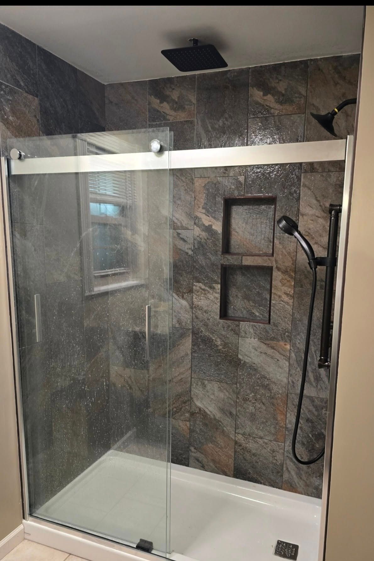 Modern shower with glass doors, dark stone tile walls, and a handheld showerhead.