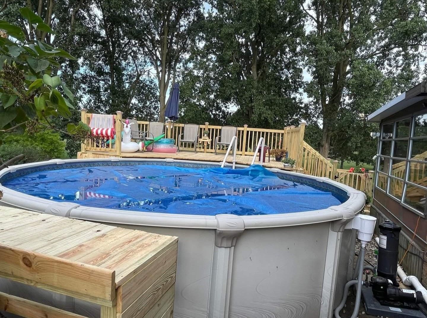 Above-ground pool with blue cover, ladder, deck chairs, and trees in a backyard