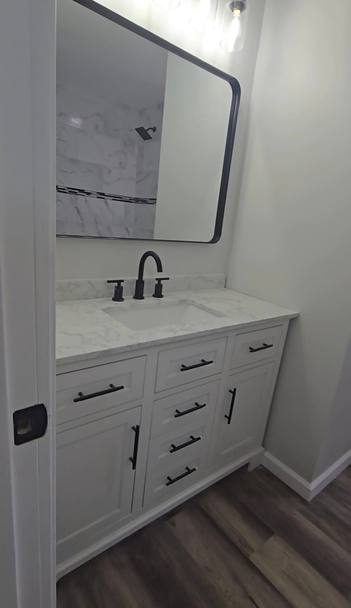 White bathroom vanity with black faucet and large mirror against light walls, wood-look floorboards.