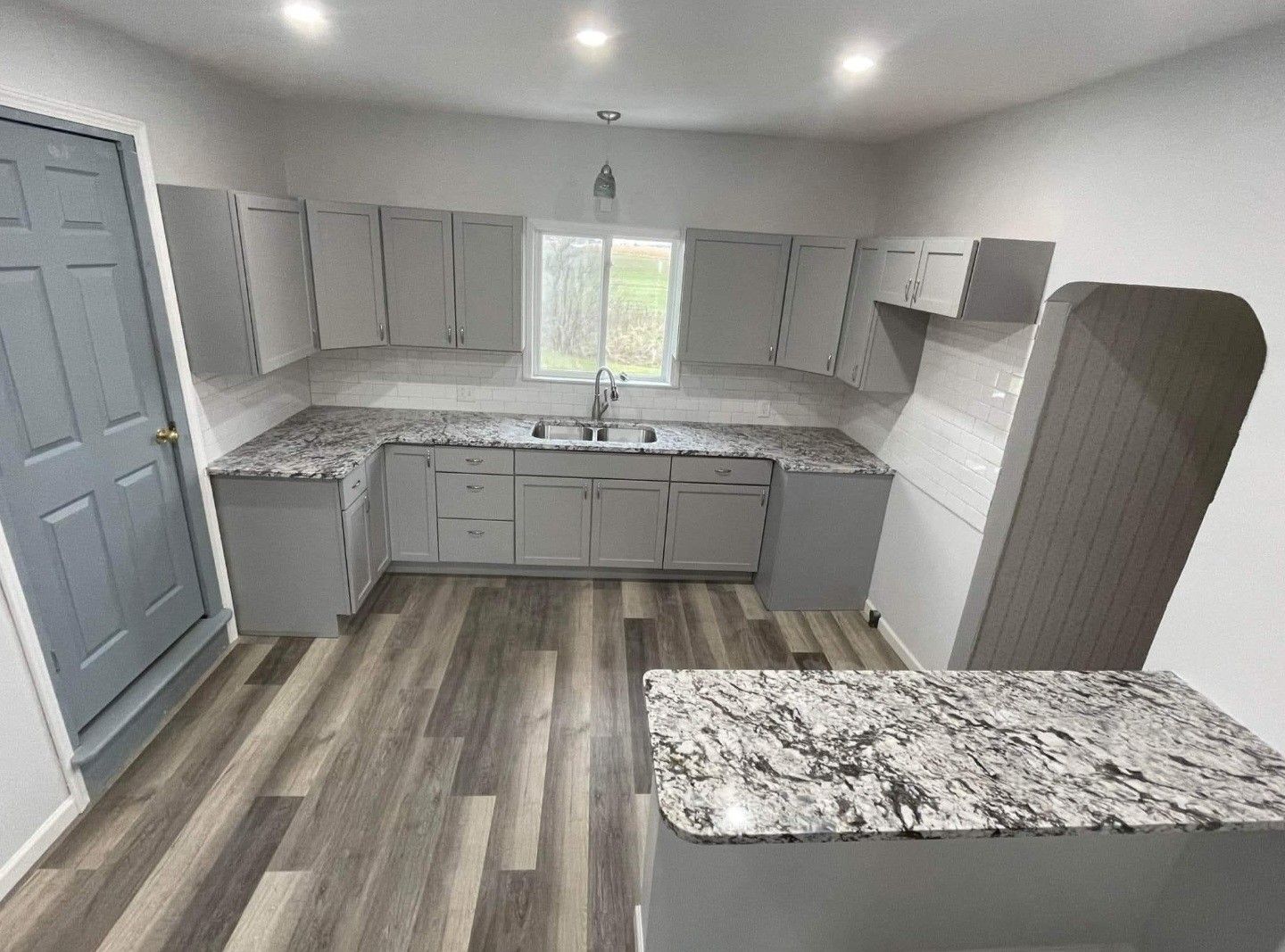 Bright modern kitchen with gray cabinets, marble counters, wood-look floor, and a window over the sink