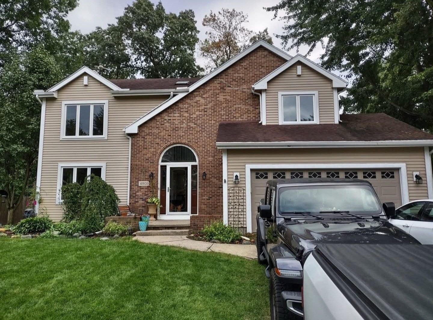 Suburban two-story house with tan siding, brick front, and two cars parked in the driveway.