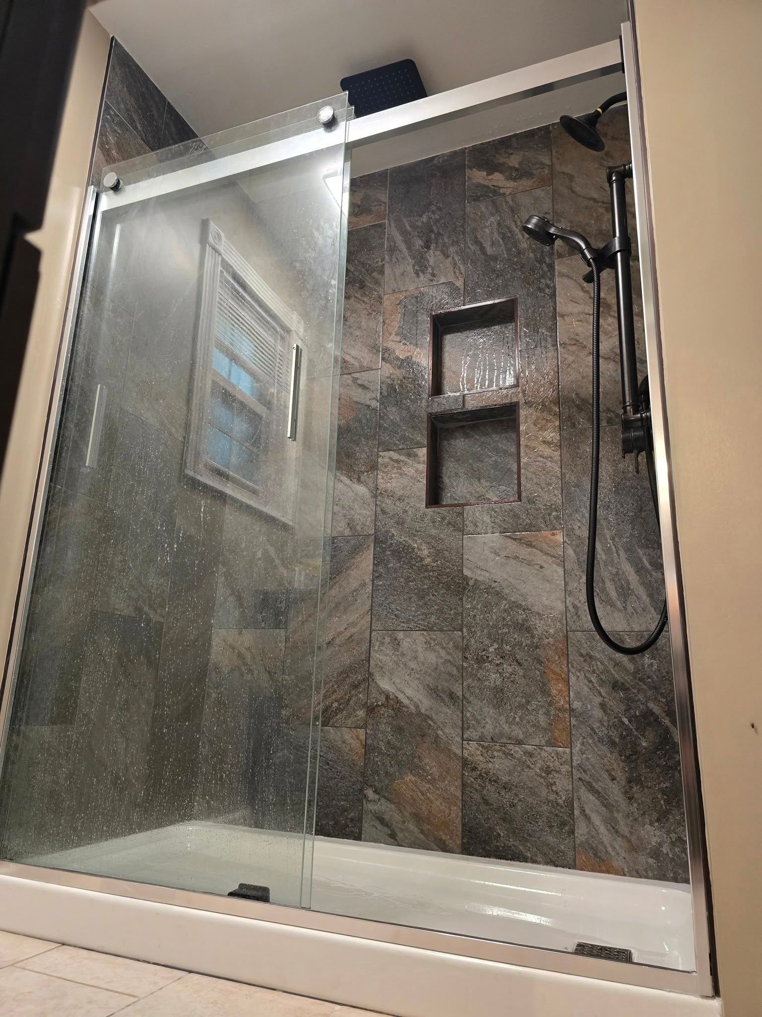 Glass shower with stone tile walls, bench, and black handheld showerhead