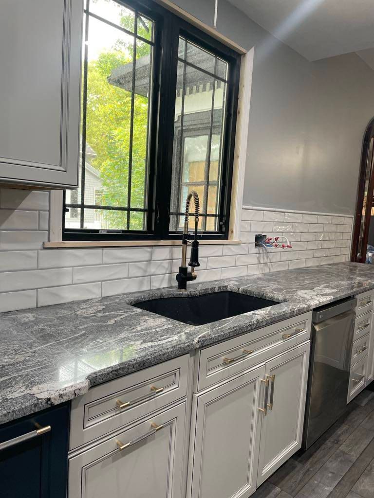 Modern kitchen sink with black faucet, marble countertop, white cabinets, and window above.