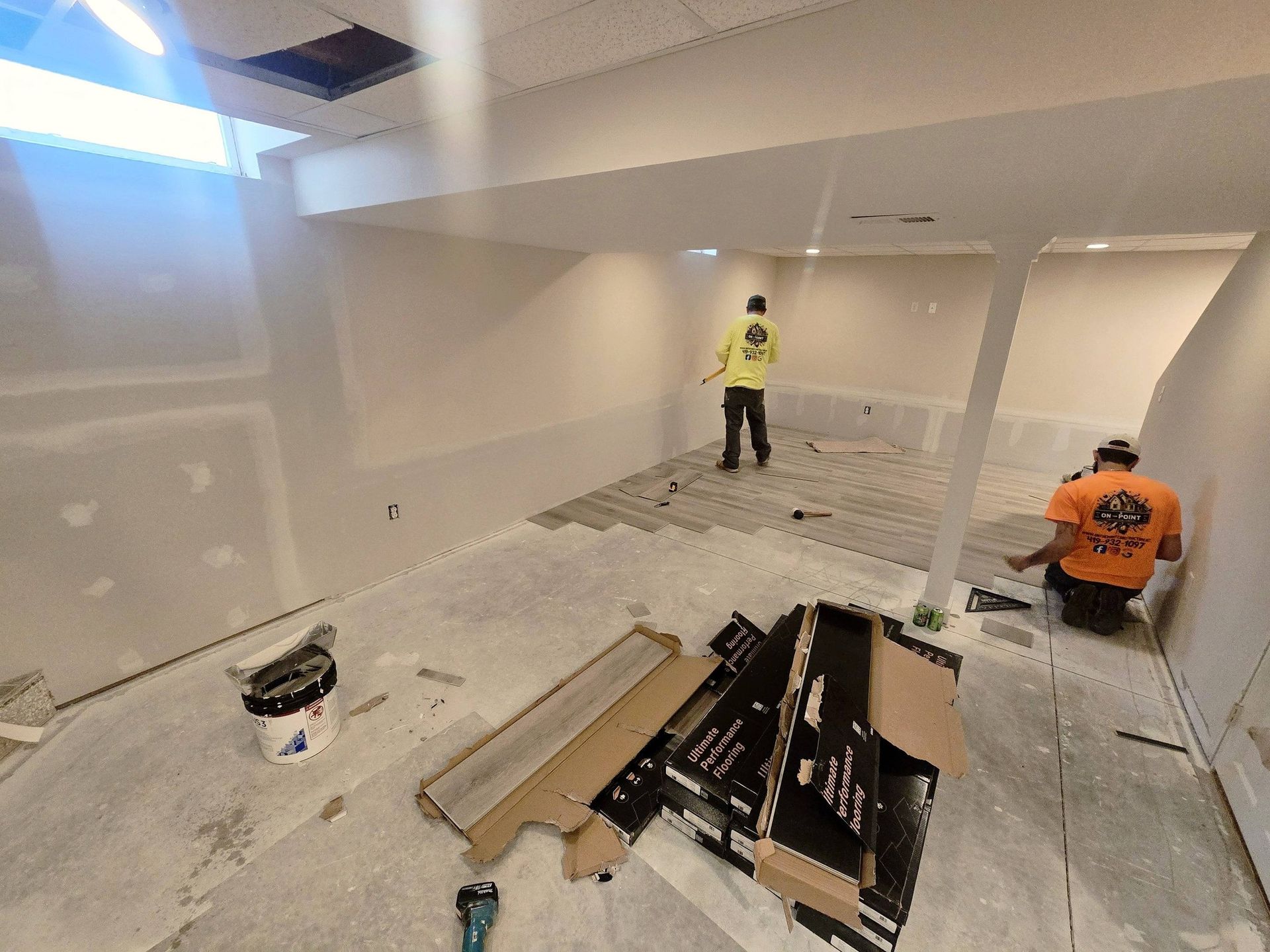 Two workers installing flooring in a bright, unfinished basement room with scattered tools and materials.
