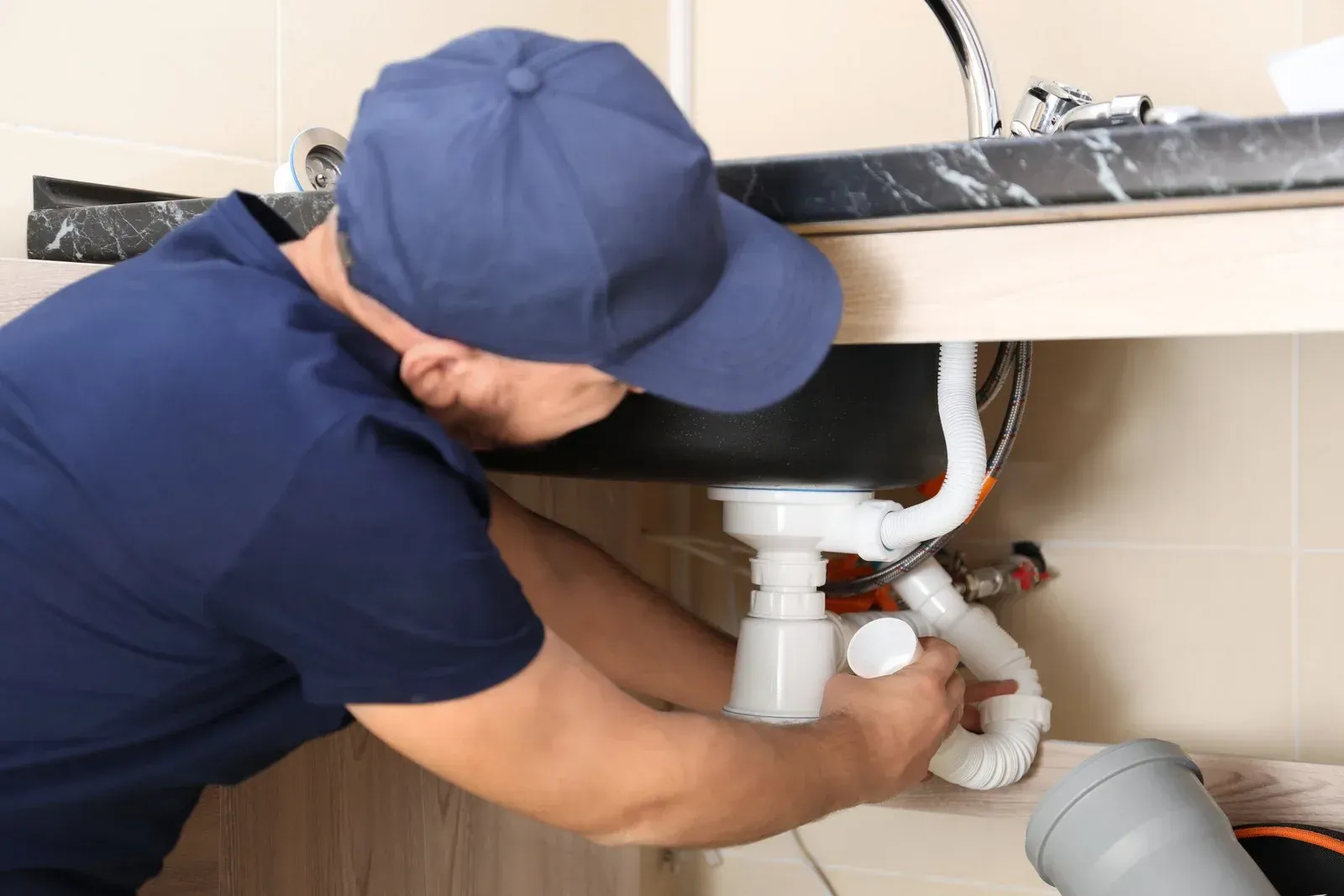 Plumber in blue uniform installing white sink plumbing under a bathroom sink.