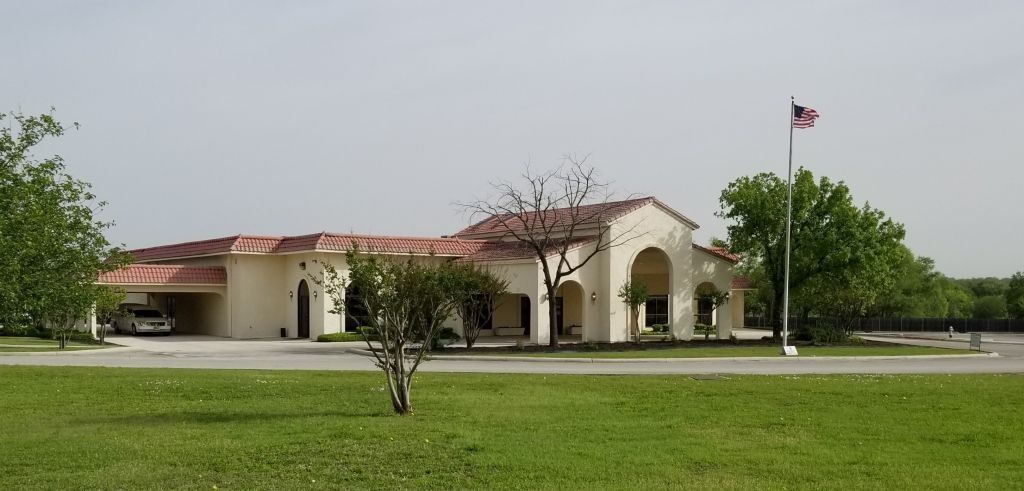 A building with a red-tiled roof and an American flag. It is surrounded by green grass and trees under a cloudy sky.
