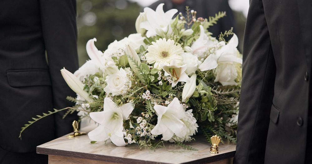 Two people in black suits carry a wooden casket with white flower arrangement.