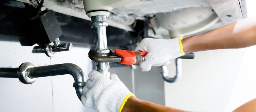 A Person Fixing Pipes Under a Sink with A Wrench, Wearing Gloves — Harwood Plumbing & Gasfitting Pty Ltd in Alawa, NT