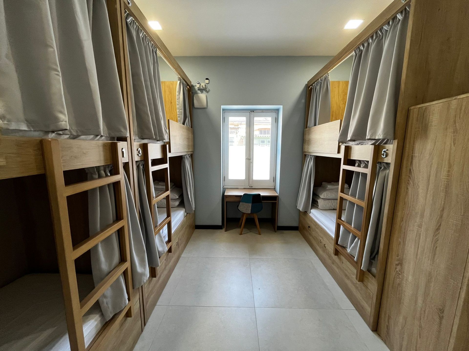 Dorm room with two rows of wooden bunk beds and a desk by a window in the center.  