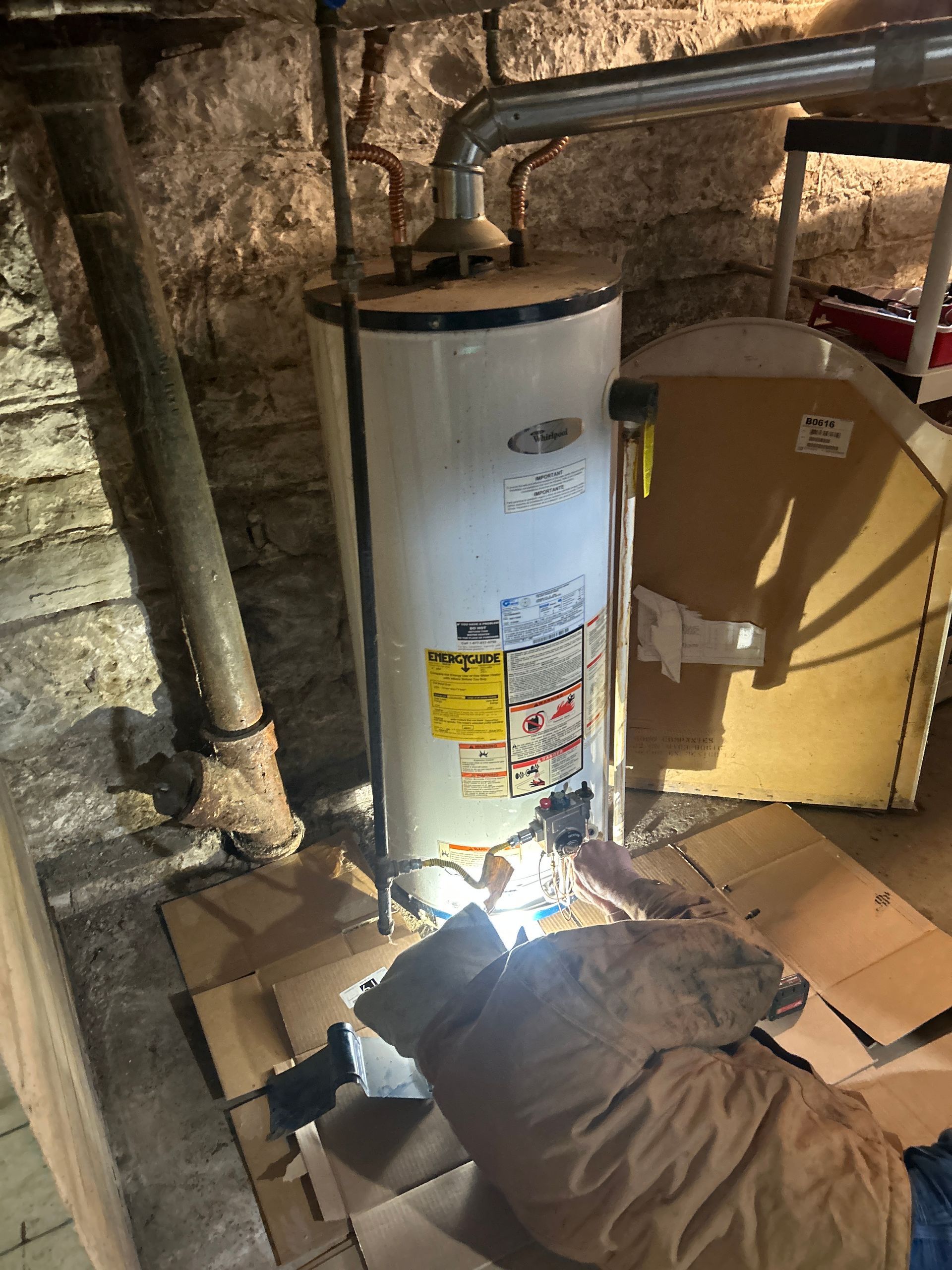 A person wearing a tan jacket kneels on cardboard to repair a white gas water heater in a stone basement.