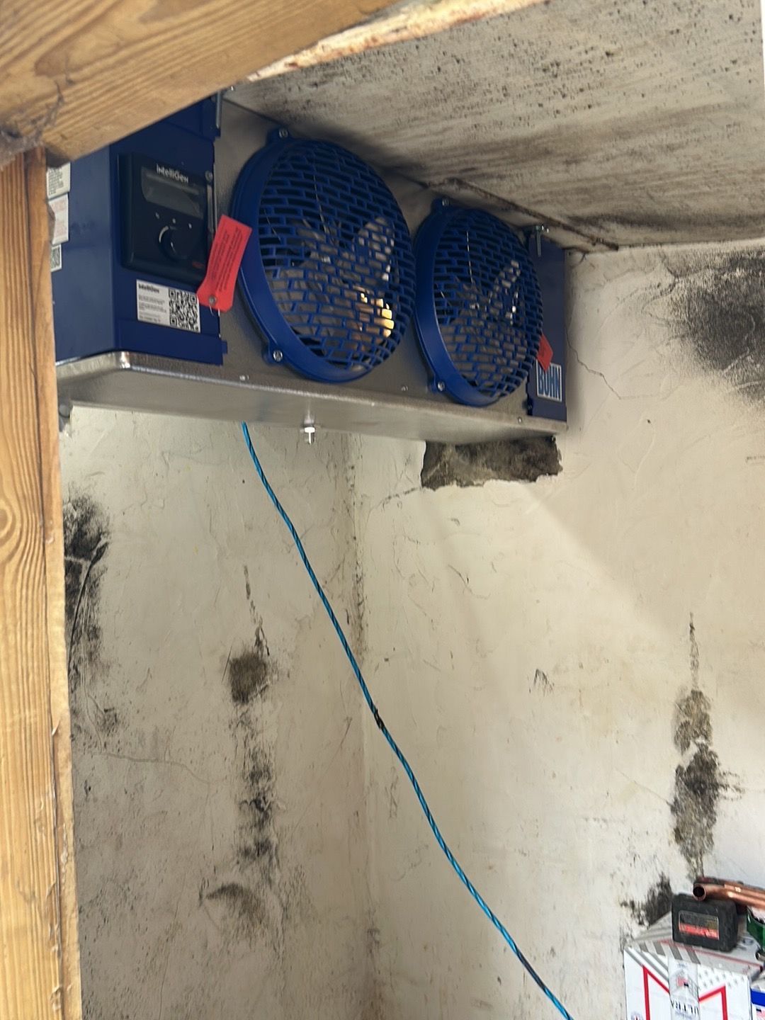 A refrigeration unit with two blue fans mounted to a concrete ceiling in a rustic interior space.