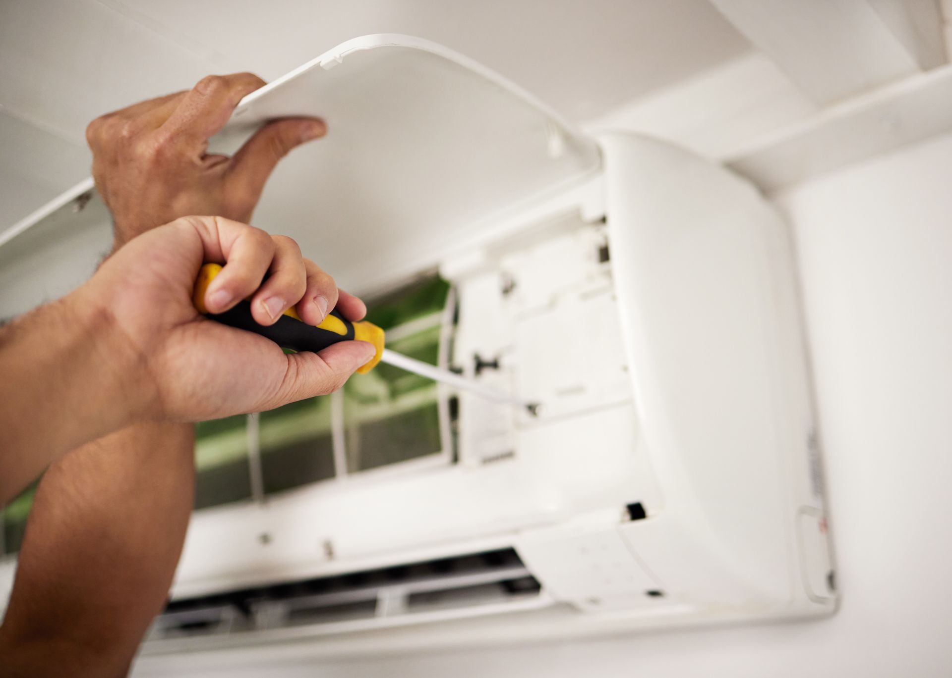 A pair of hands uses a screwdriver to service the interior components of a white wall-mounted air conditioning unit.