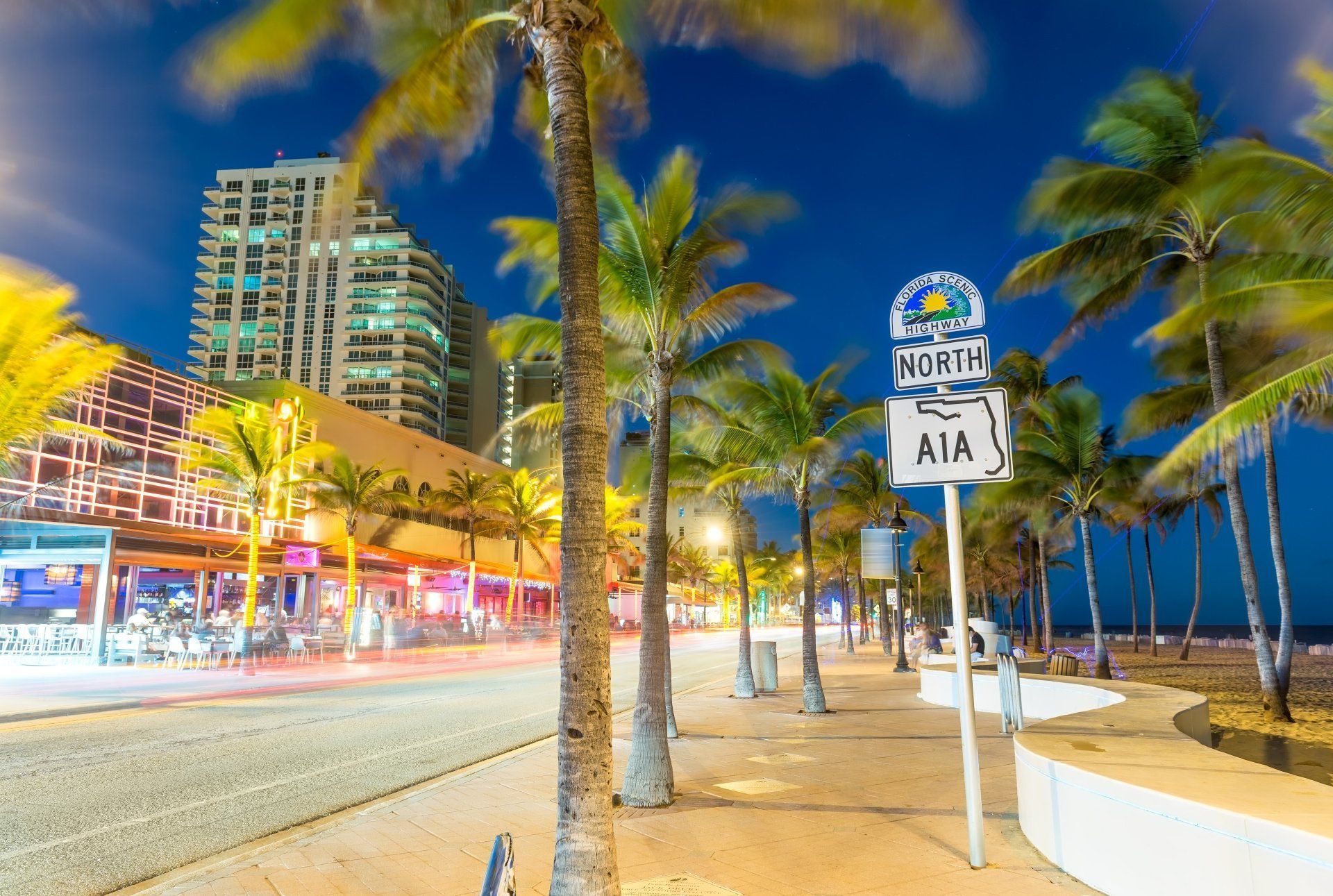 Nighttime beach boulevard with palm trees, lit buildings, and a North A1A sign by the shoreline.