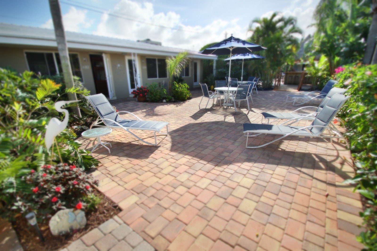 Patio with brick pavers, outdoor chairs, round table, and umbrella beside a home with tropical plants