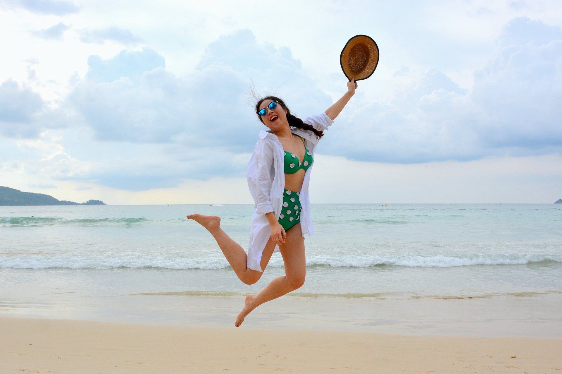 Woman in green swimsuit jumping on a beach, waving a hat by the ocean under a bright sky