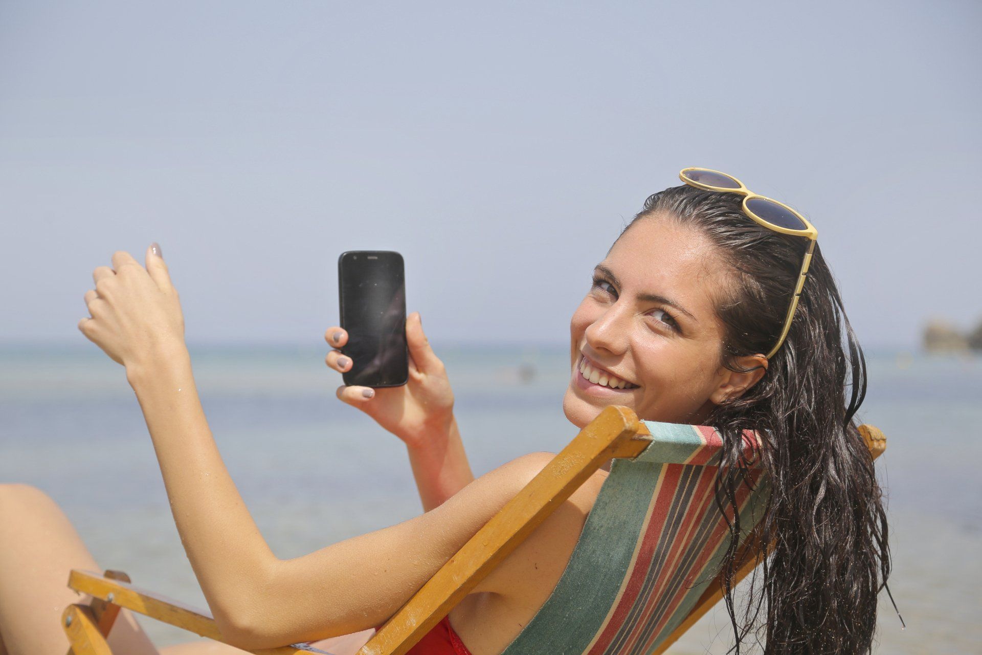 Woman smiling on a beach chair, holding a phone and waving toward the ocean.