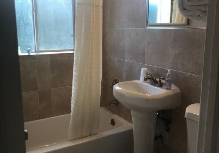 Small bathroom with a bathtub, pedestal sink, toilet, and beige tiled walls near a window.