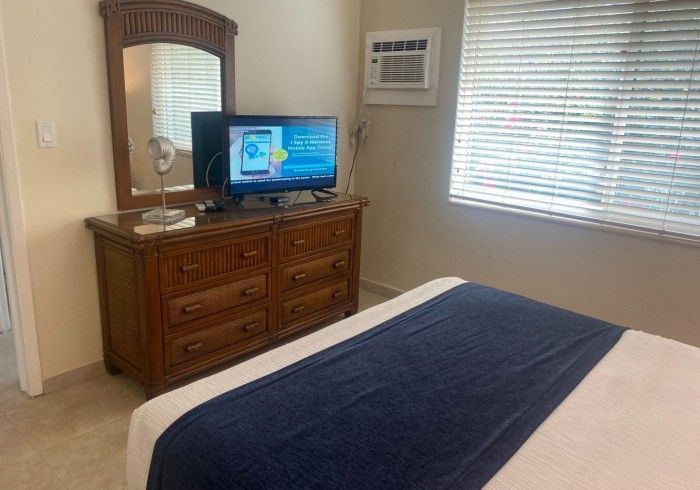 Bedroom with a wooden dresser, TV, wall mirror, and a bed with a blue runner.