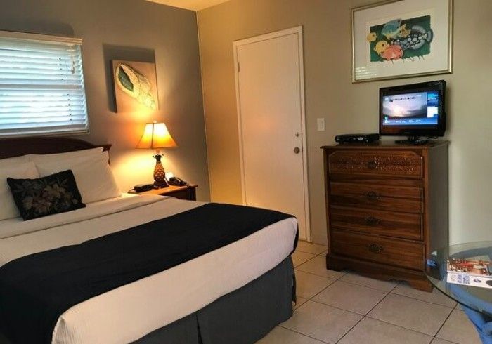Small bedroom with a bed, lamp, dresser, TV, and wall art on beige walls.