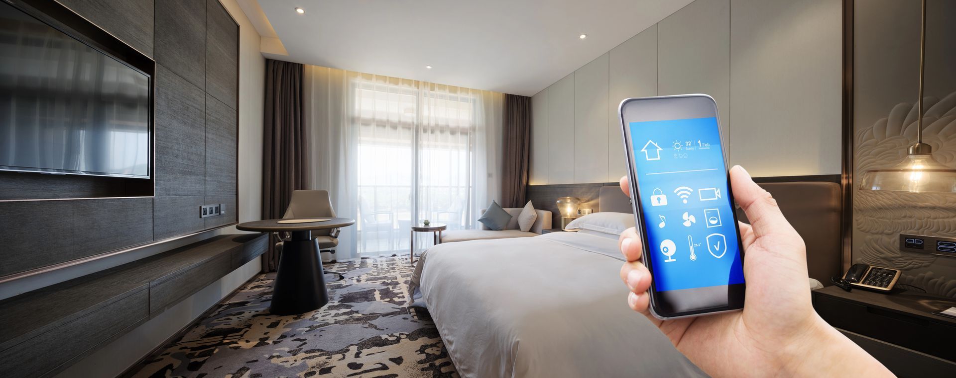Una mano tiene un smartphone en una moderna camera d’albergo con un letto, una scrivania e una grande finestra.