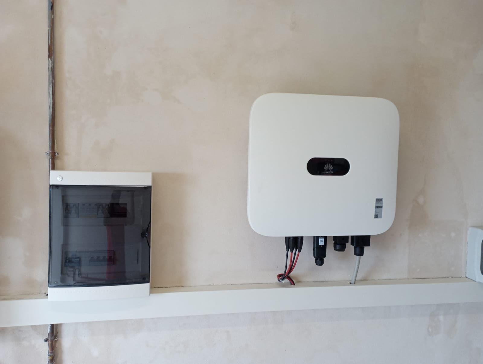 Dispositivo bianco a parete con cavi sopra una mensola, accanto a un piccolo quadro elettrico su una parete beige